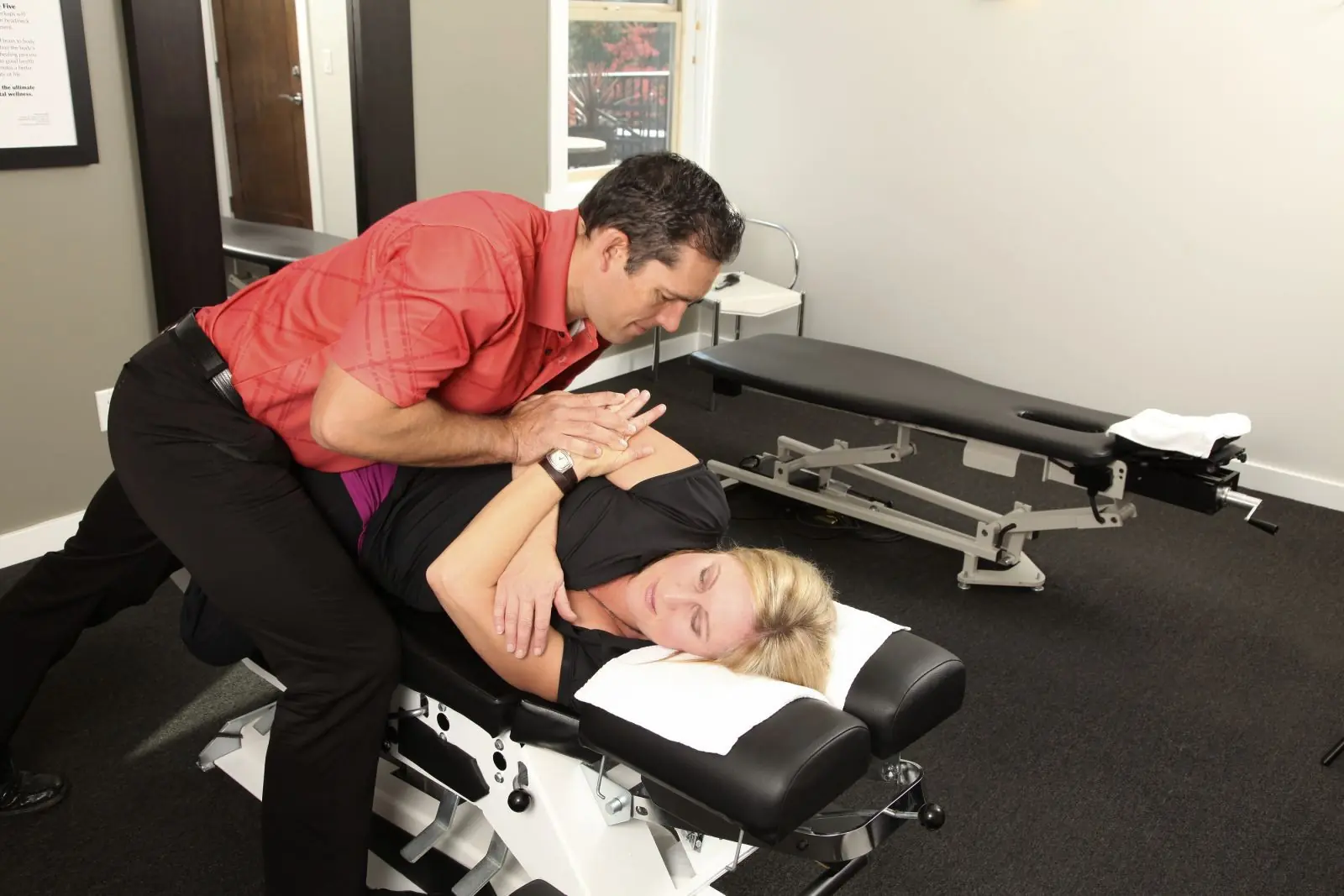 Chiropractor in red shirt performing an adjustment on a woman lying face down on a treatment table.