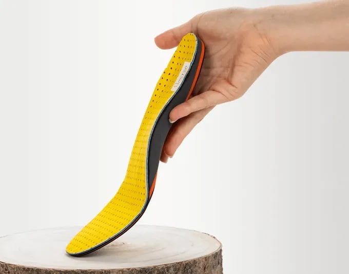 Hand holding a yellow and black shoe insole above a wooden stump against a white background.