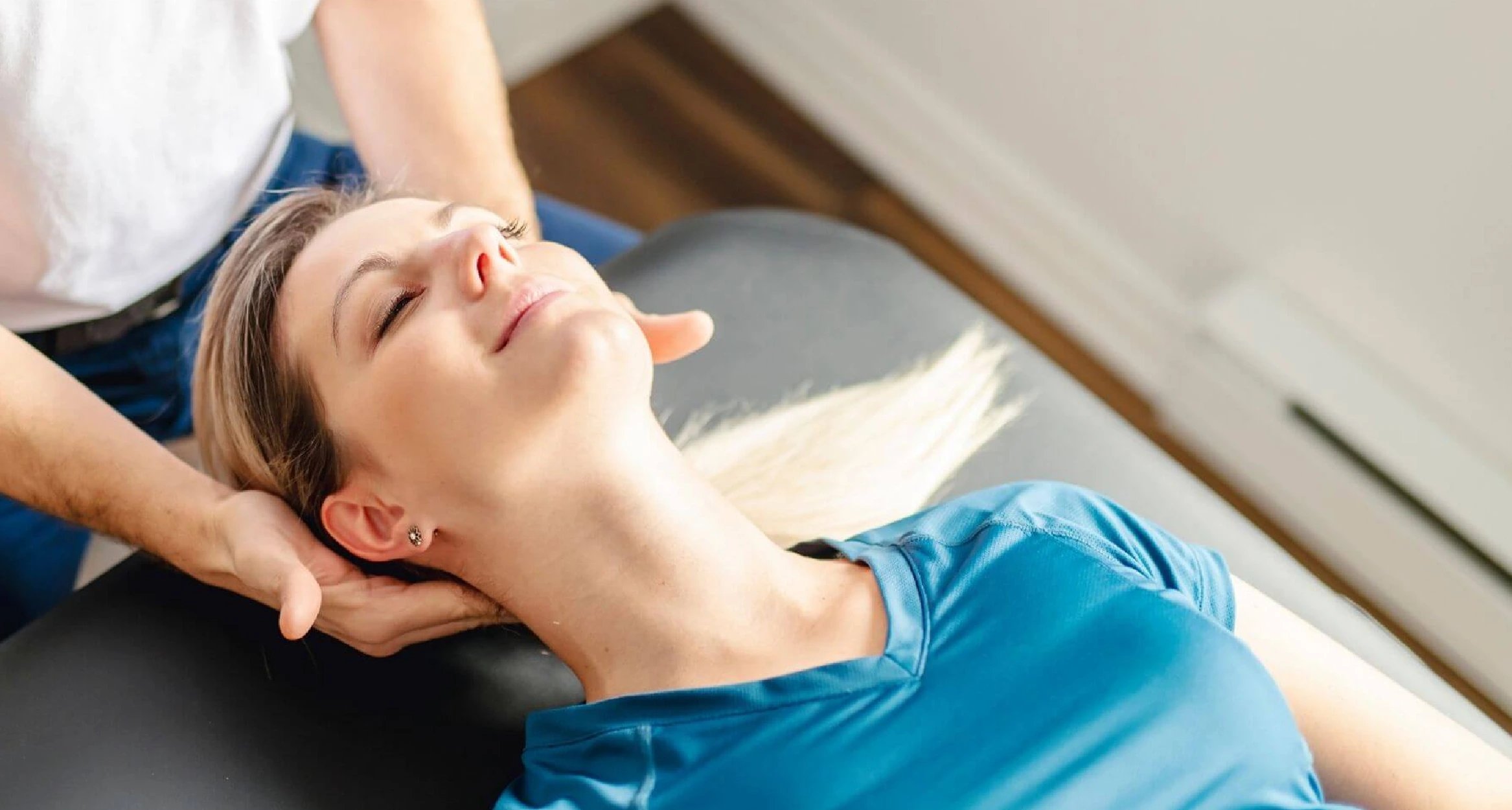 A woman in a blue shirt relaxes as a person gently supports her head during a massage or therapy session.