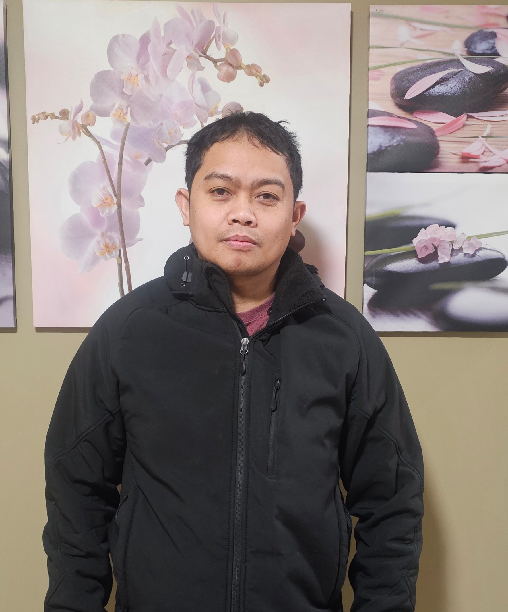 Man in black jacket standing indoors against wall with orchid and stone art panels.