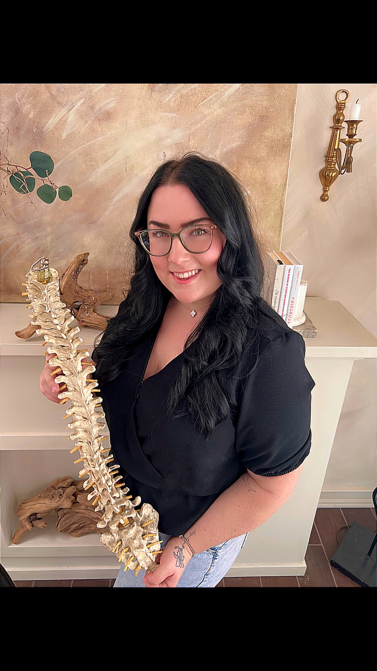 Smiling woman with glasses holds a spine model in a room with books and wall decor.