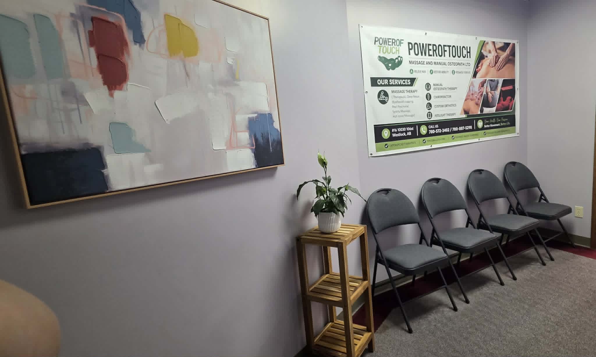 Waiting area with four gray chairs, a plant on a wooden stand, abstract art, and a massage service sign.