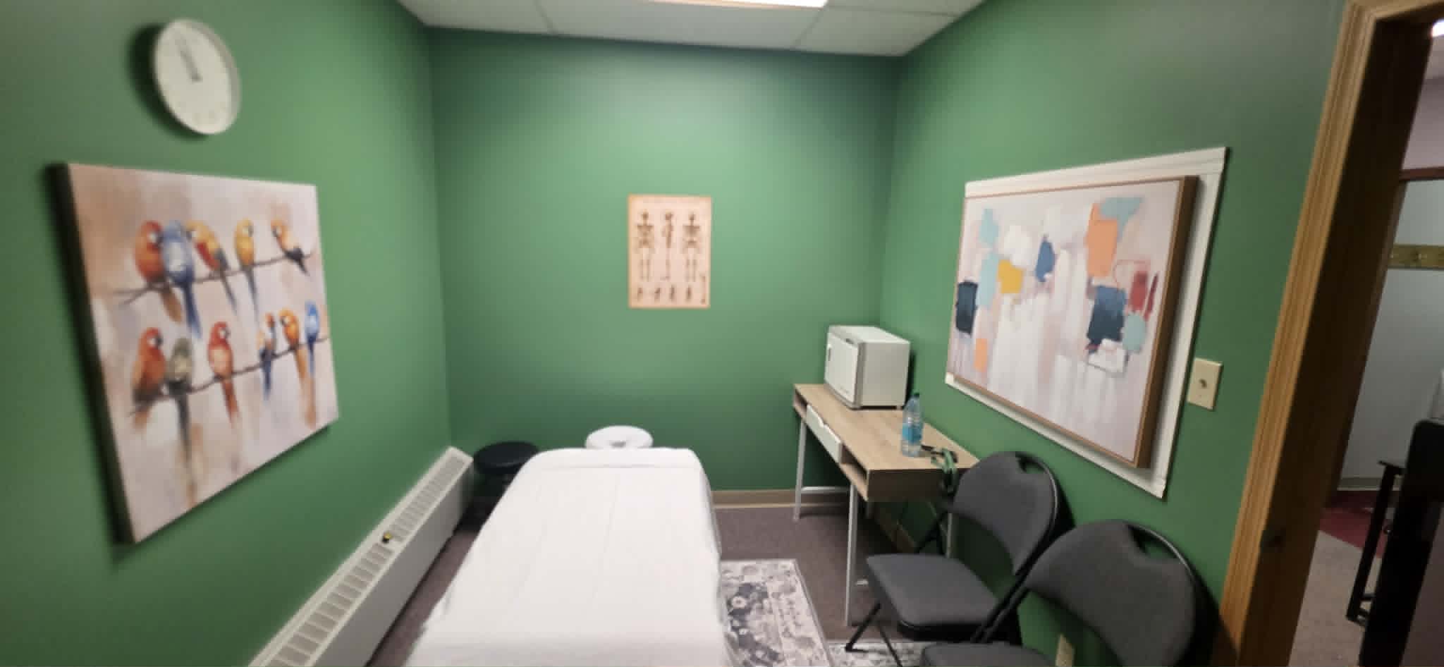 Small green massage room with white table, two chairs, colorful bird and abstract paintings.