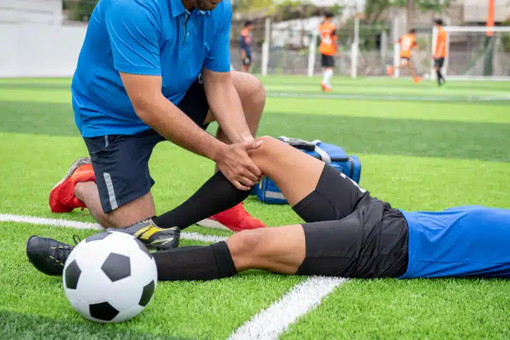 Person in blue shirt kneeling on grass, examining injured soccer player's knee during a game.
